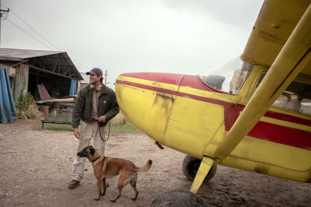 Elokuva-arvostelu: The Dog Stars on Ridley Scottin parhaita elokuvia vuosiin, mutta sillä on surullinen kohtalo Jacob Elordi ävelee koiran kanssa keltaisen pienlentokoneen ja lentokonehallin vieressä The Dog Stars -elokuvassa.