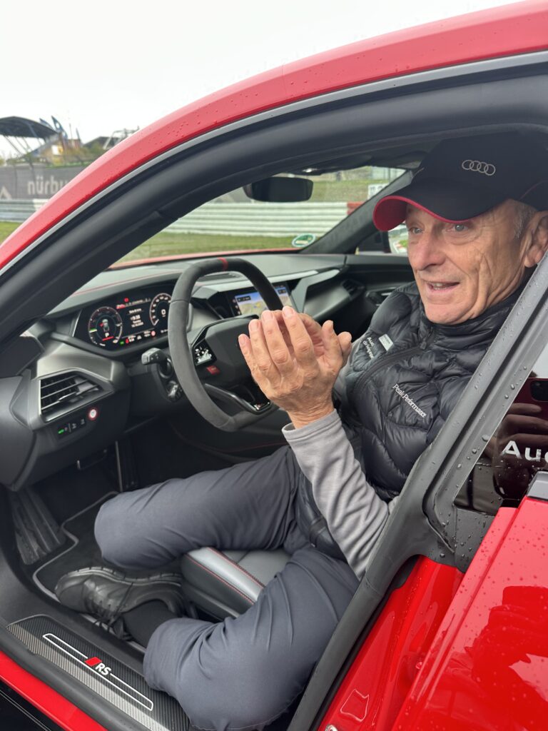 Justsiks koki Audi driving experiencen Nürburgringillä: märkää rataa ja unohtumaton ajopäivä Harald Büttner istuu punaisen Audin kuljettajan paikalla ja elehtii käsillään avoimen sivuikkunan ääressä.