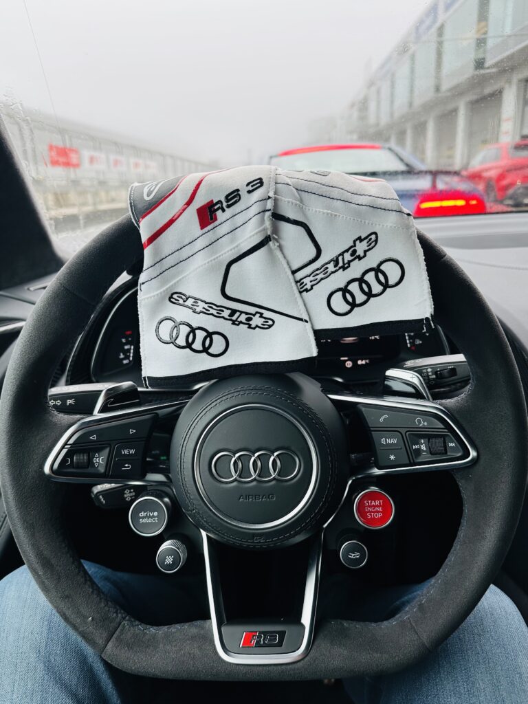 Justsiks koki Audi driving experiencen Nürburgringillä: märkää rataa ja unohtumaton ajopäivä Valkoiset Audi-ajohanskat R8 ohjauspyörällä ja autoja sateisella varikkosuoralla.